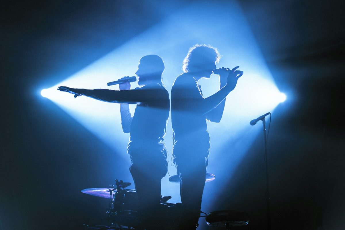 Two singers stand back to back in a dark room with spotlights shining on each one