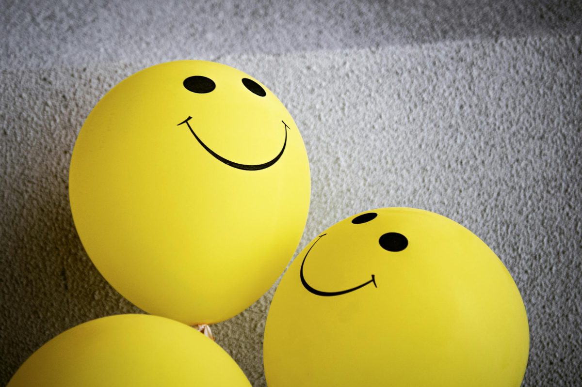 Yellow balloons with smiley faces on them sit in a pile against a white wall